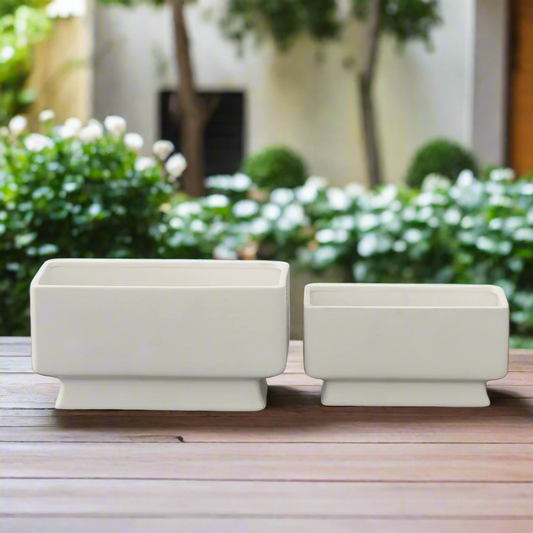 Two matte white ceramic rectangular pots with curved edges and flared bottom base, set on a wooden surface with foliage in the background.