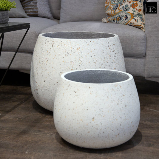 Two white cement flower pots with a speckled finish, one larger than the other, placed on a floor next to a piece of furniture.